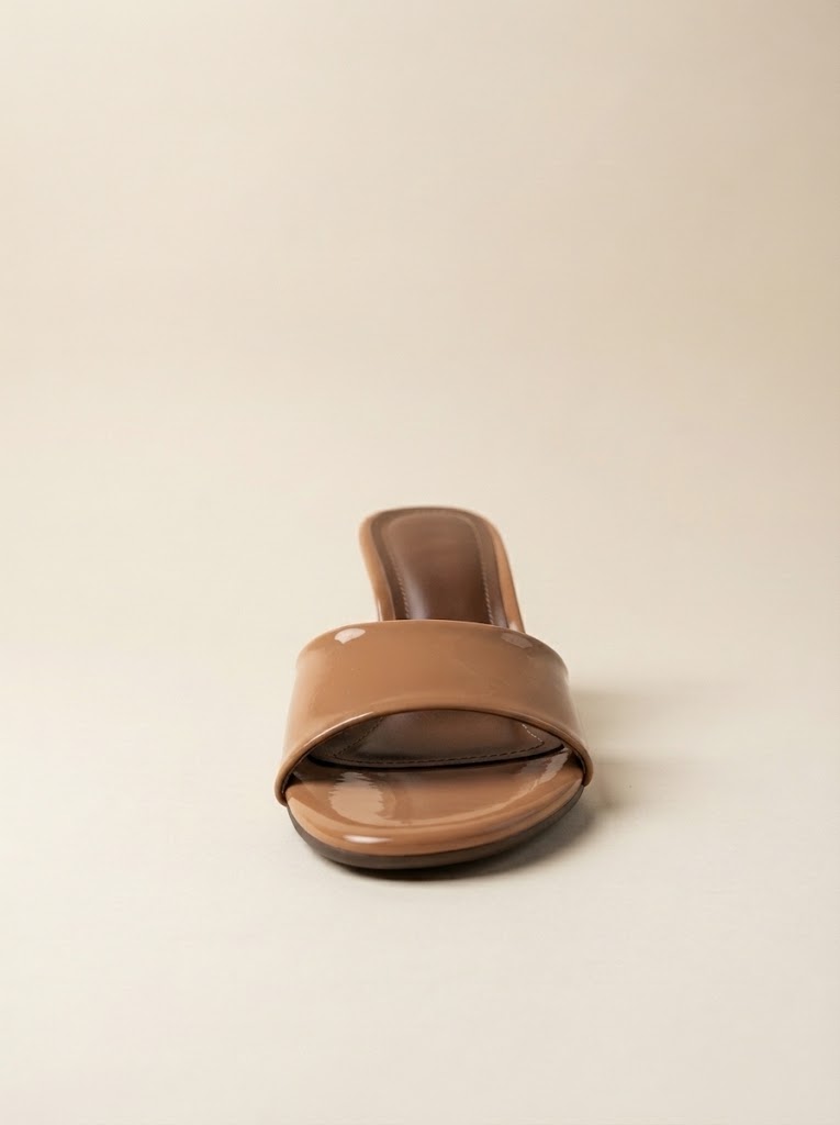 A single tan patent leather slide sandal with a low block heel, shown from a front-facing perspective on a neutral beige background.