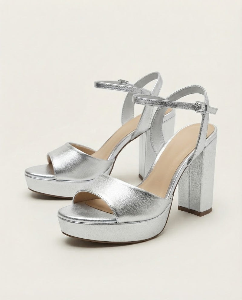 A pair of metallic silver platform high-heeled sandals with ankle straps, shown on a light beige studio background.