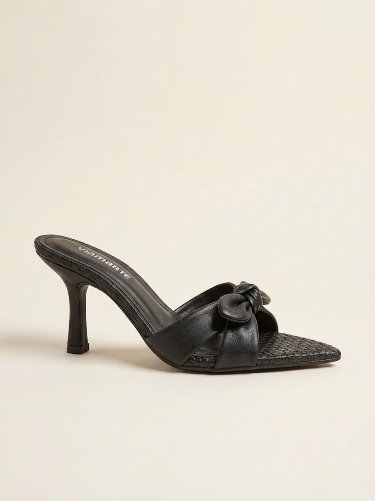 A single black leather high-heeled mule sandal with a pointed toe, textured woven pattern on the toe box, and a decorative bow detail on the instep, shown against a plain light beige background.