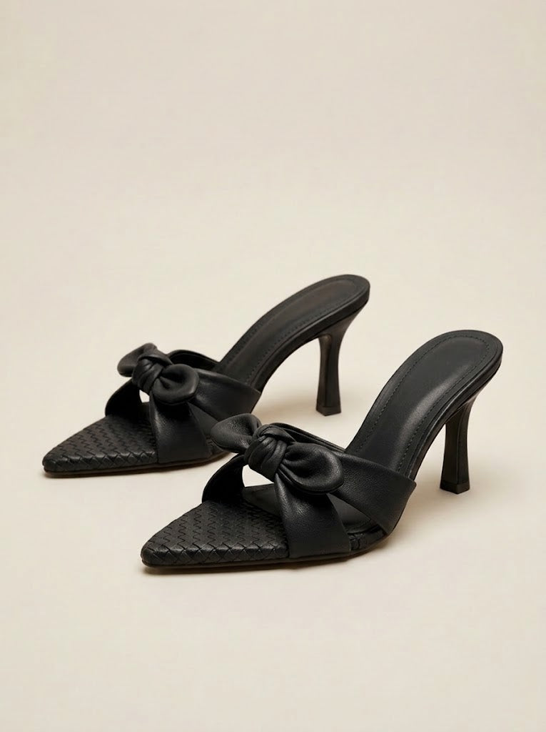 A pair of black pointed-toe mule heels with a woven texture on the toe strap, a wide satin bow detail across the instep, and slim kitten heels, shown on a neutral beige background.