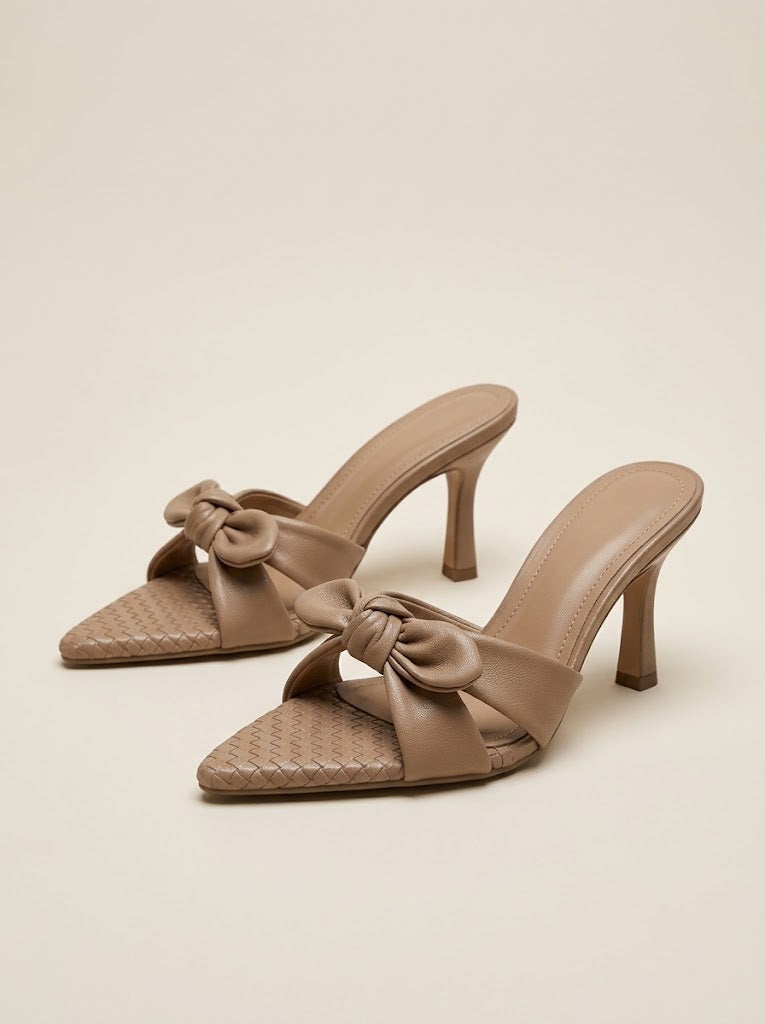 A pair of taupe pointed-toe mule heels with woven detailing on the toe and a large satin bow across the instep, set against a solid beige background.