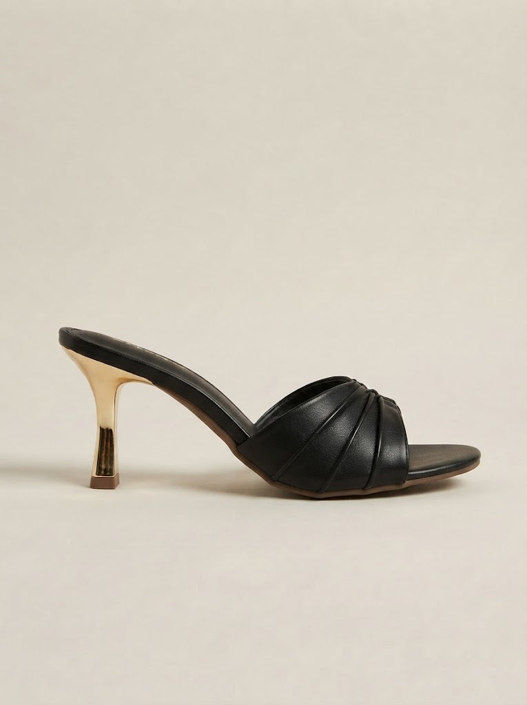 A single black leather mule sandal with a crisscross strap design and a slim metallic gold high heel, shown against a plain beige background.