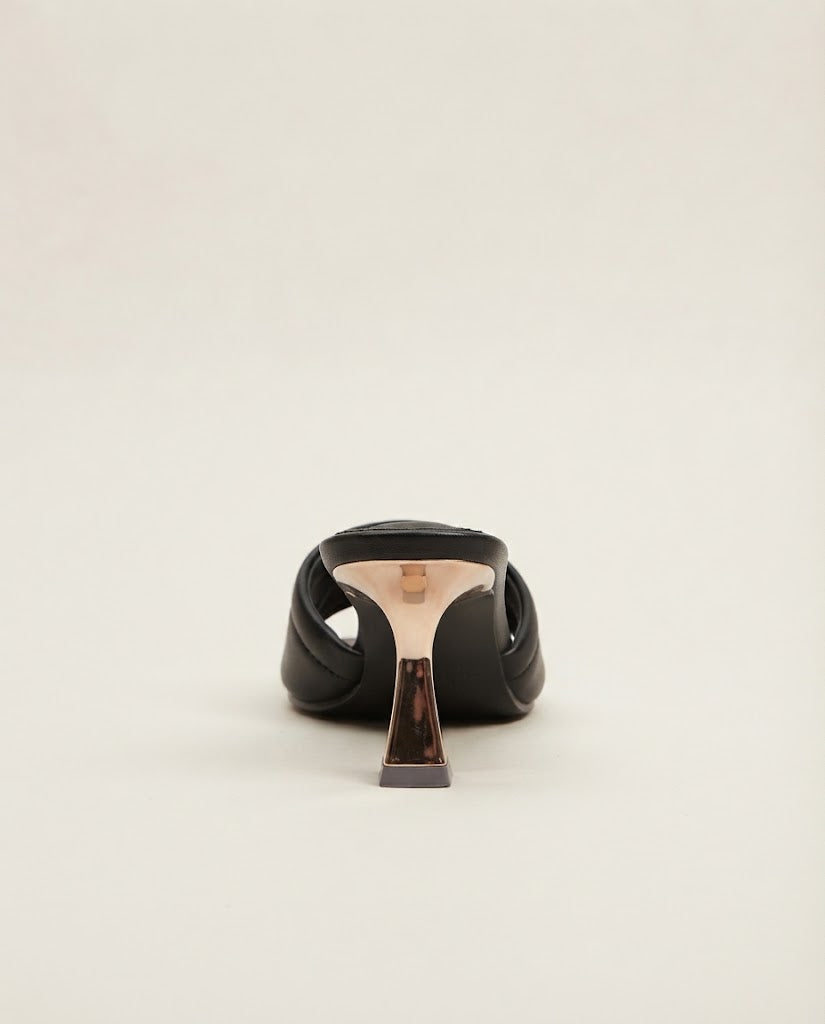 A single black leather high-heeled sandal shown from the back, featuring a sculptural metallic rose gold heel and a dark brown tortoiseshell accent block, set against a plain off-white background.