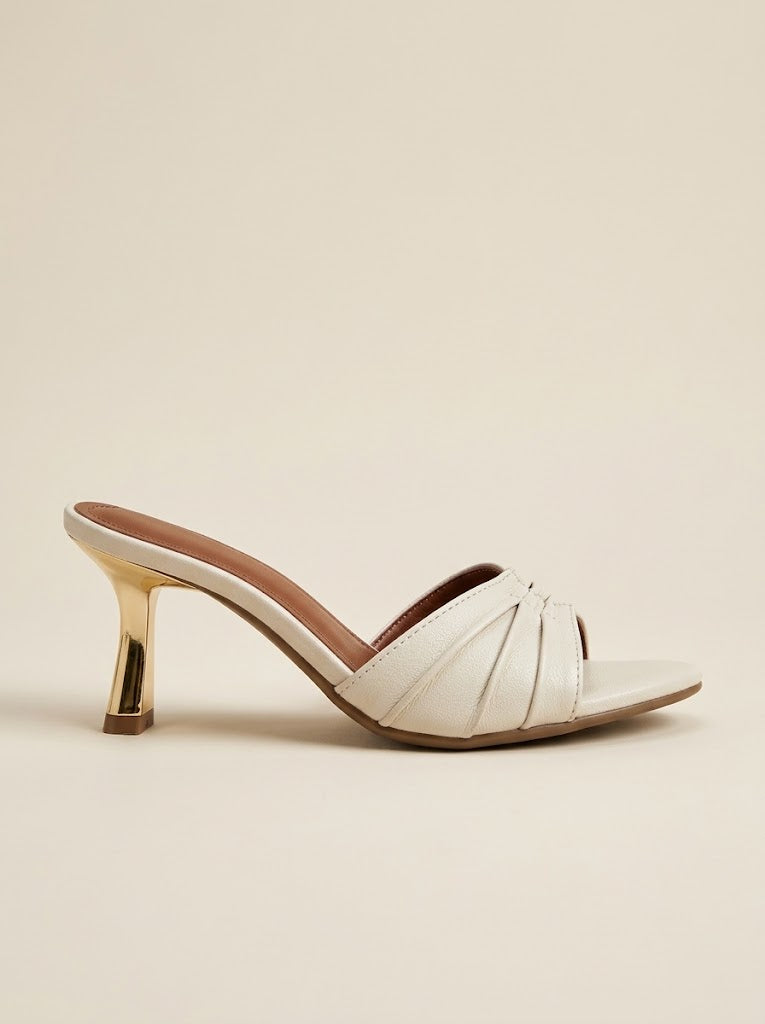 A single white leather mule sandal with a pointed toe, crisscross strap design, and a metallic gold block heel, shown against a solid beige background.
