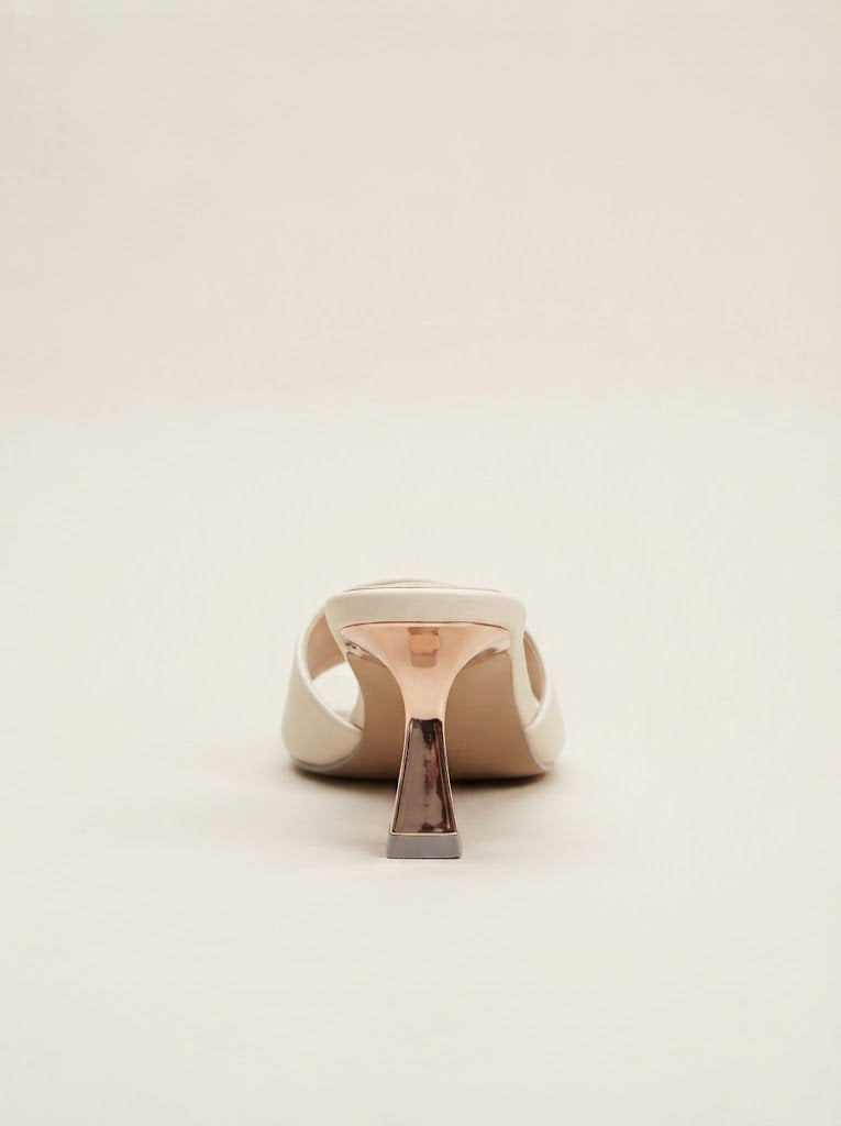 A single beige leather mule sandal shown from the back, featuring a sculptural metallic rose gold and silver geometric heel on a solid cream background.
