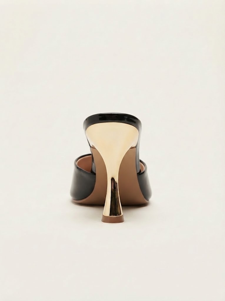 A single black leather mule heel with a sculptural metallic gold block heel and a tan interior, shown from a rear three-quarter angle on a light cream background.