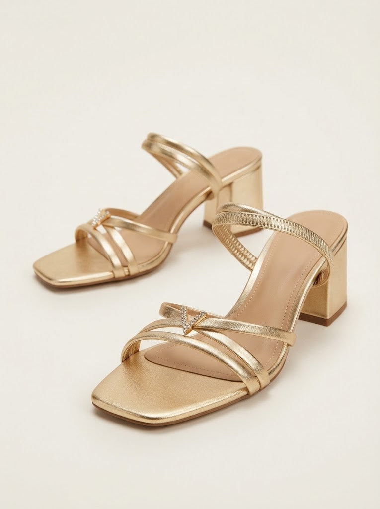 A pair of metallic gold strappy mule sandals with square toes and block heels, displayed on a light beige background.