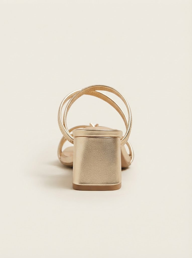 A single metallic gold block-heel sandal with thin wrap-around straps, shown from a rear three-quarter angle on a clean, light beige background.