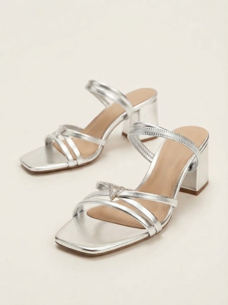 A pair of metallic silver strappy mule sandals with square toes and block heels, displayed on a light beige background.