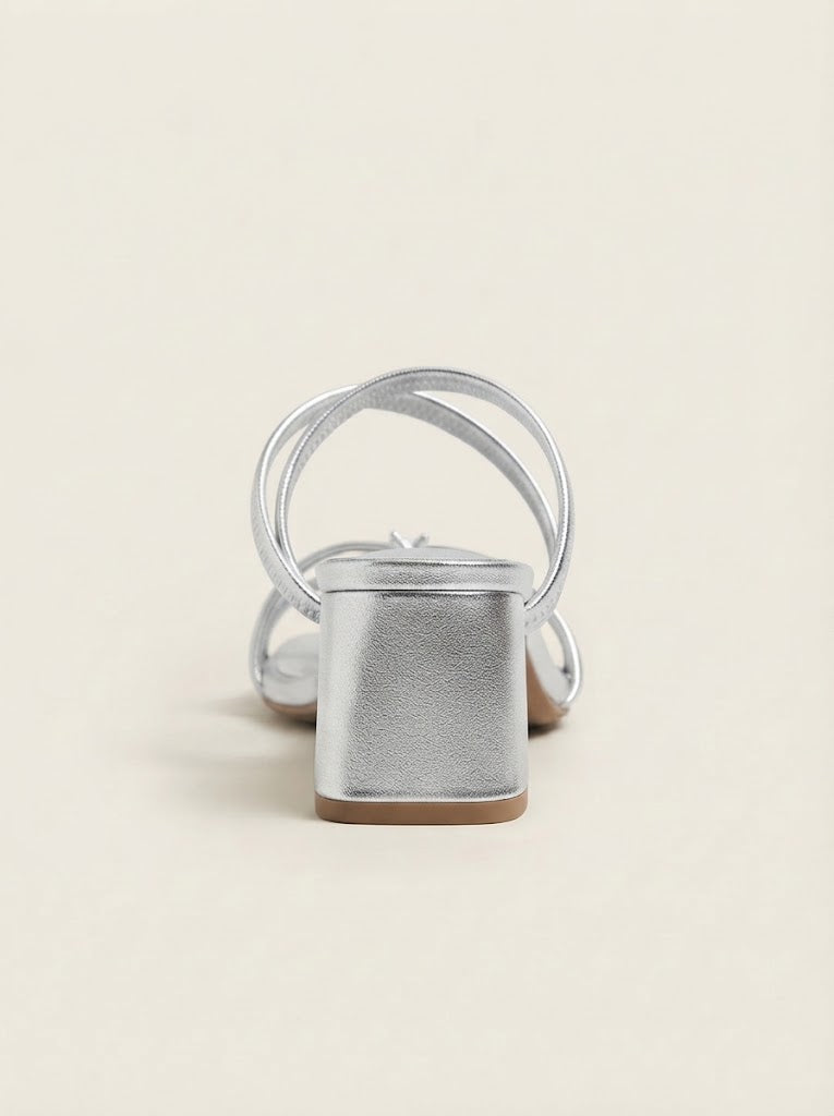A single metallic silver block-heel sandal with thin wrap-around straps, shown from a rear three-quarter angle on a light beige background.