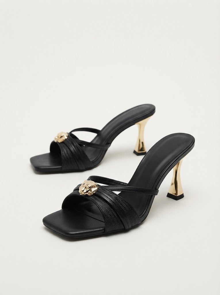 A pair of black leather mule sandals with square toes, textured straps, a gold-tone decorative metal accent, and a slim gold-tone high heel, shown on a light gray studio background.
