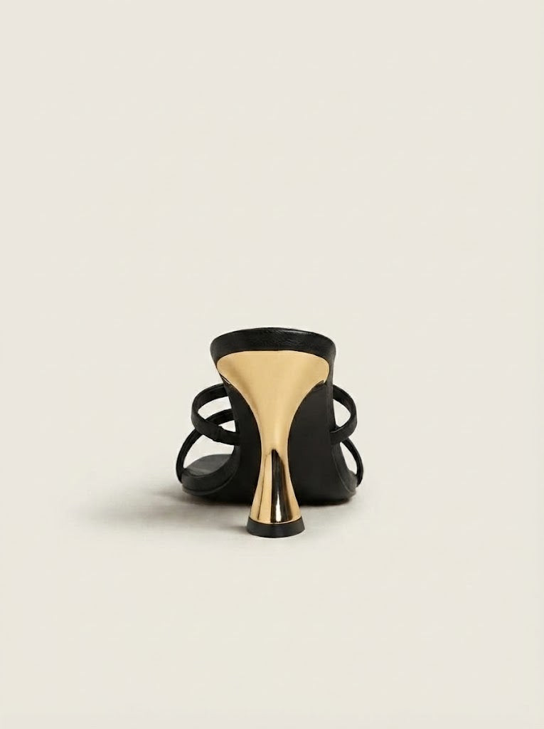 Black strappy high-heeled sandals with a unique metallic gold conical heel, shown from a rear three-quarter angle on a light beige background.