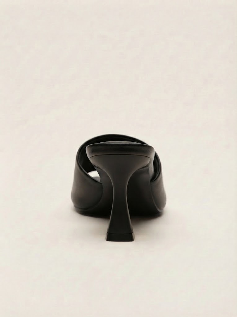 A single black leather mule sandal shown from the back, featuring a chunky mid-height stiletto heel and a wrap-around strap design, set against a plain light beige background.