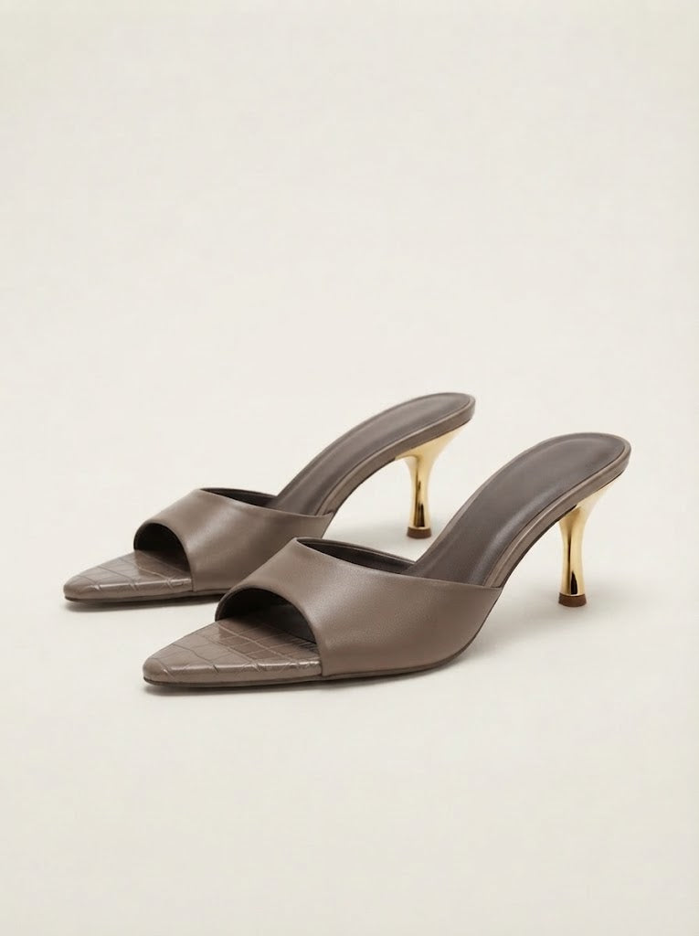 A pair of taupe leather mule heels with pointed toes, a subtle crocodile-embossed pattern on the toe, and slim gold stiletto heels, set against a plain light beige background.