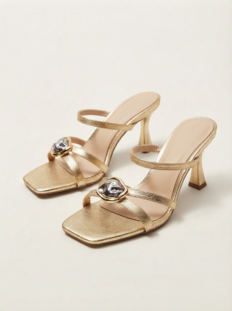 A pair of metallic gold heeled sandals with square toes, thin straps, and heart-shaped crystal embellishments on the straps, shown on a light beige background.