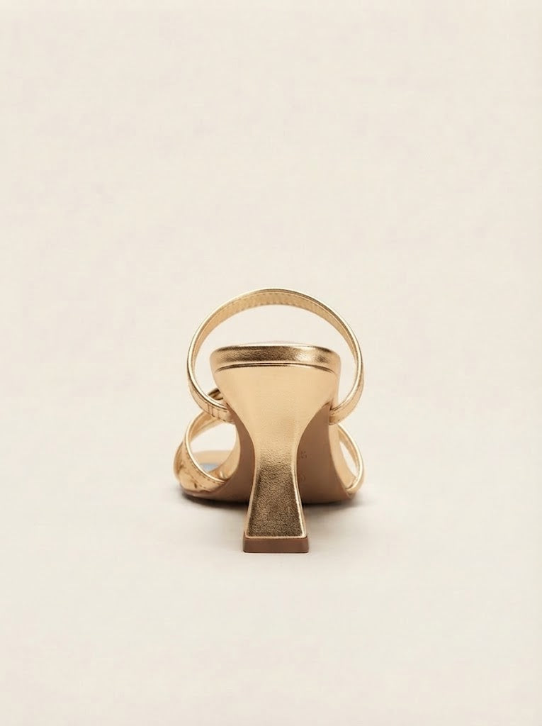 A single metallic gold high-heeled sandal shown from the back on a plain, warm off-white background.