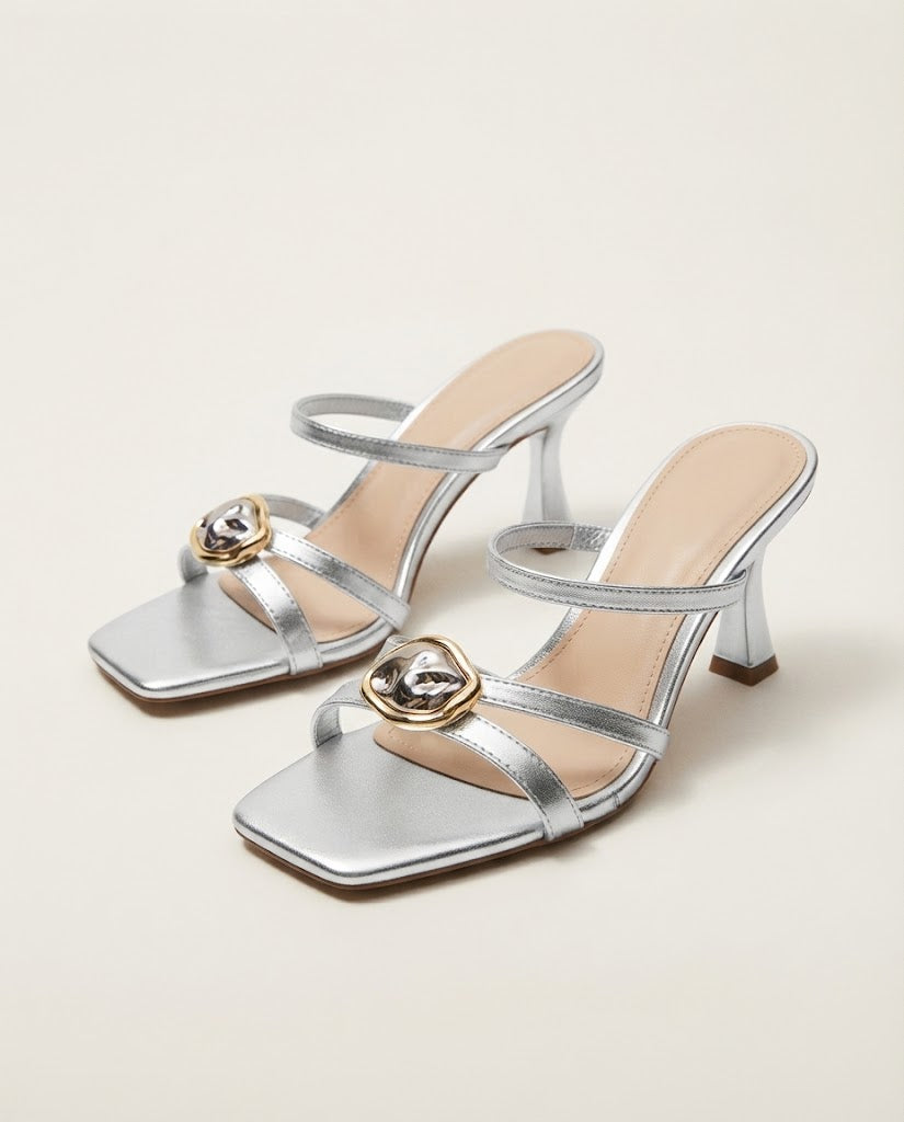 A pair of metallic silver heeled mule sandals with square toes, thin straps, and a decorative gold-tone heart-shaped embellishment on the front, shown on a light beige background.