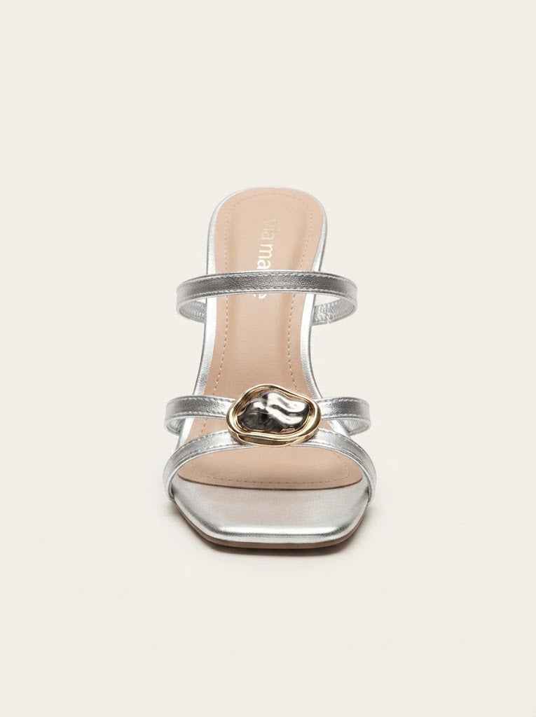 A single metallic silver heeled sandal with a square toe, thin straps, and a central gold-tone heart-shaped embellishment, shown against a plain off-white background.